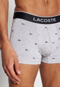 Grey boxer briefs made of soft fabric, featuring a black waistband with "LACOSTE" branding and small black logo patterns throughout.