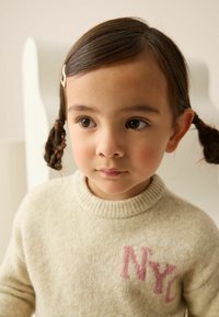 Cream-colored knit sweater with ribbed collar and pink "NYC" text. Child with brown hair in pigtails, wearing a hair clip. Soft texture.