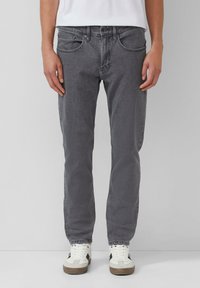 Persoon in slim-fit grijze spijkerbroek en witte sneakers, staand tegen een effen lichtgrijze achtergrond.