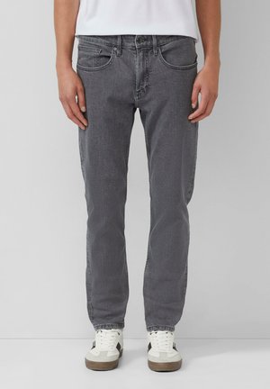 Persoon in slim-fit grijze spijkerbroek en witte sneakers, staand tegen een effen lichtgrijze achtergrond.