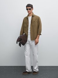 Braunes Leinenhemd über einem weißen T-Shirt, kombiniert mit weißen Hosen und braunen Wildlederschuhen. Hält eine dunkelbraune Tasche und trägt eine dunkle Sonnenbrille.