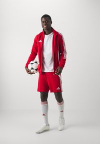 Rød atletisk tracksuit med hvide Adidas striber, hvid t-shirt, røde shorts og hvide fodboldstøvler. Holder en sort og hvid fodbold.