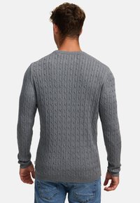 Grå kabelstrikket genser med rund hals, ribbestrikkede mansjetter og teksturert stoffmønster, iført over blå jeans sett fra baksiden.