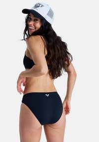Ensemble bikini noir avec un haut ajusté et un bas taille basse. Le modèle porte une casquette en maille blanche avec le logo "ROXY" en noir. Texture lisse, design simple.