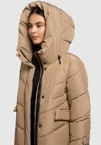 Béžová péřová bunda s přehnaně velkou kapucí, vodorovnou prošívanou strukturou, předním zipem a dvěma bočními kapsami. Hladká látka s jemným leskem.