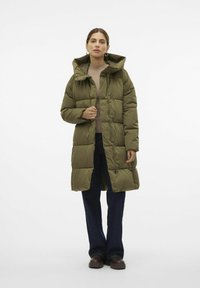 Abrigo acolchado de color verde oliva con capucha, que presenta un diseño acolchado, cierre frontal de cremallera y bolsillos. Combinado con jeans de campana de color azul oscuro.