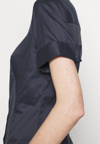 Mörkblå kortärmad skjorta med en slät, glänsande yta, med uppvikta ärmslut och en knappdetalj på ärmen.