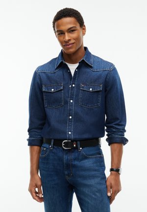 Young man wearing a dark blue denim shirt with two chest pockets, blue jeans, a black belt, and a black watch standing against a white background.