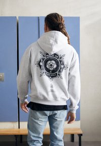 Hoodie gris avec un design graphique noir au dos, incluant un lion et du texte. Manches longues, poche kangourou, matériau en mélange de coton.