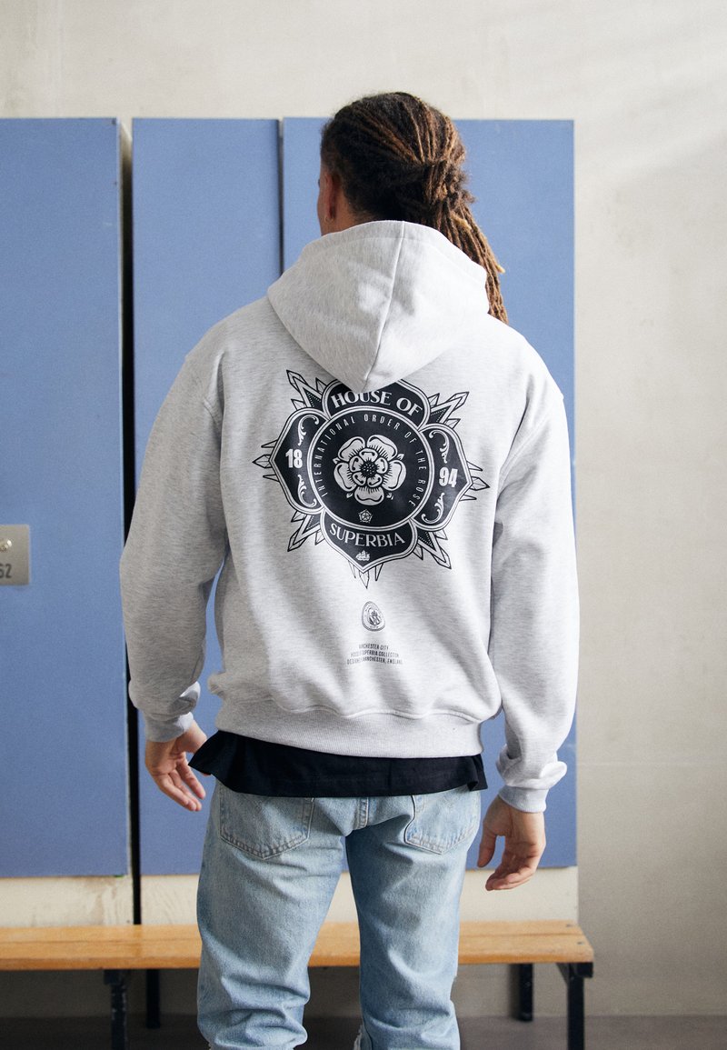 Hoodie gris avec un design graphique noir au dos, incluant un lion et du texte. Manches longues, poche kangourou, matériau en mélange de coton.