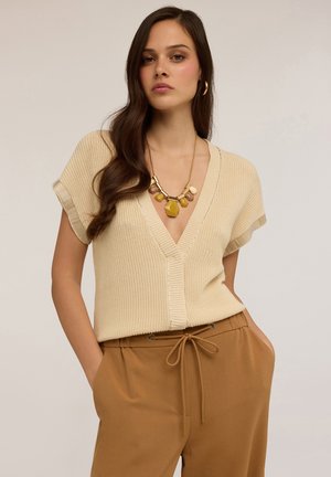 Beige Strickoberteil mit tiefem V-Ausschnitt und kurzen Ärmeln, kombiniert mit braunen Hosen mit Gummizug. Goldkette als Akzent.