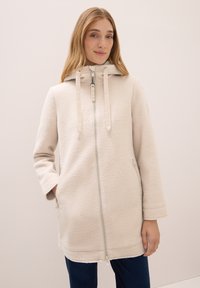 Donna che indossa un cappotto con cappuccio di un beige chiaro e texturizzato, con una zip frontale e un cordoncino, in pose con una mano in tasca su uno sfondo semplice.