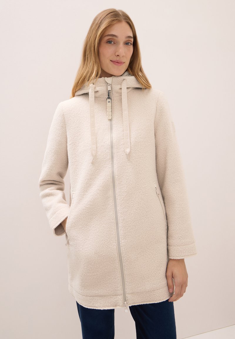 Donna che indossa un cappotto con cappuccio di un beige chiaro e texturizzato, con una zip frontale e un cordoncino, in pose con una mano in tasca su uno sfondo semplice.