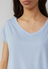 Niebieski, krótki top z rękawem, z dekoltem w kształcie litery V. Materiał wydaje się gładki i lekki, co podkreśla luźny krój. Minimalne detale przeszycia.