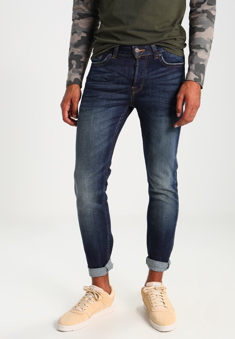 Man som bär mörkblå uppvikta jeans, långärmad camouflage-tröja, olivgrön skjorta och beige sneakers står på vit bakgrund.