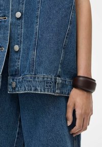 Denim jaka ar sudraba spiedpogām un izšūtu logo, kombinēta ar gludu tumši brūnu rokassprādzi uz rokas, izceļot slāņotu izskatu.