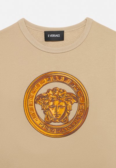 Pull beige avec un emblème brodé doré de tête de Méduse centré sous l'encolure ronde, étiquette noire avec le texte « E VERSACE ».