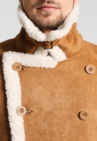Abrigo de ante marrón con un cuello blanco peludo, que presenta un diseño diagonal, acentos de botones y un detalle de hebilla visible en el cuello.