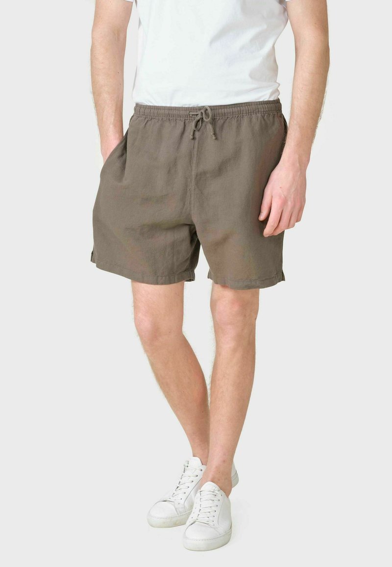 Homme portant un short marron à cordon, un t-shirt blanc et des baskets blanches, debout avec les mains dans les poches sur un fond uni.