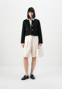 Veste noire courte avec boutons dorés, short plissé crème et mocassins noirs. Un petit sac à main blanc complète la tenue.
