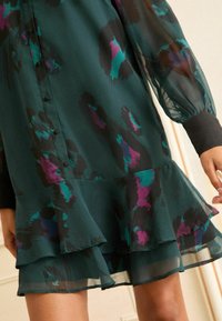 Rochie florală din voal de culoare verde închis, cu motive negre și violet. Poate fi observat un tiv ondulat, mâneci lungi cu puff și detalii cu închidere prin butoane.