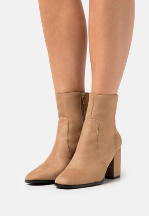 Botines de ante marrón con tacones cuadrados y cremalleras laterales, llevados sobre piernas desnudas frente a un fondo blanco.