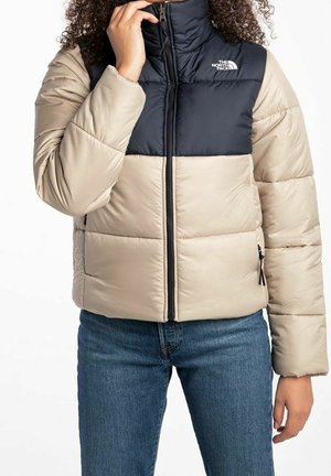 Person iført beige og mørkeblå dunjakke med lynlås, trækker kraven op, kombineret med blå denimjeans.