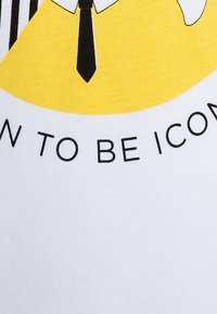 Camiseta blanca con un gráfico amarillo que presenta una ilustración de una corbata y un texto negro en negrita que dice "NACIDO PARA SER ICÓNICO." Textura de algodón suave.