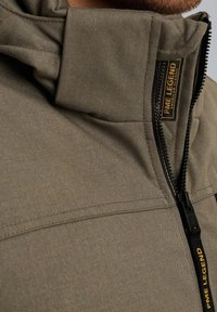 Khaki kurtka z wysokim kołnierzem, teksturowany materiał i widoczny czarny zamek. Posiada metkę z logo na klatce piersiowej oraz wyraźnie zarysowane szwy.