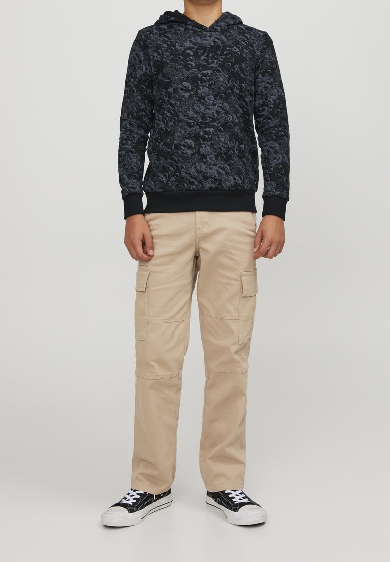 Personne portant un sweat à capuche noir avec un motif crâne, un pantalon cargo beige et des baskets noires et blanches, se tenant devant un fond uni.