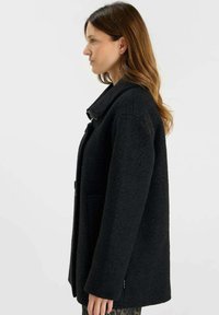 Zwarte wollen jas met een gestructureerde kraag, zijzakken en een losse pasvorm. De textuur lijkt zacht en lichtjes gestructureerd, en biedt warmte.