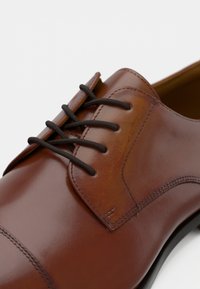 Chaussure habillée en cuir marron avec une finition lisse, bout rond et cinq paires de lacets marron foncé. Couture détaillée le long des coutures.