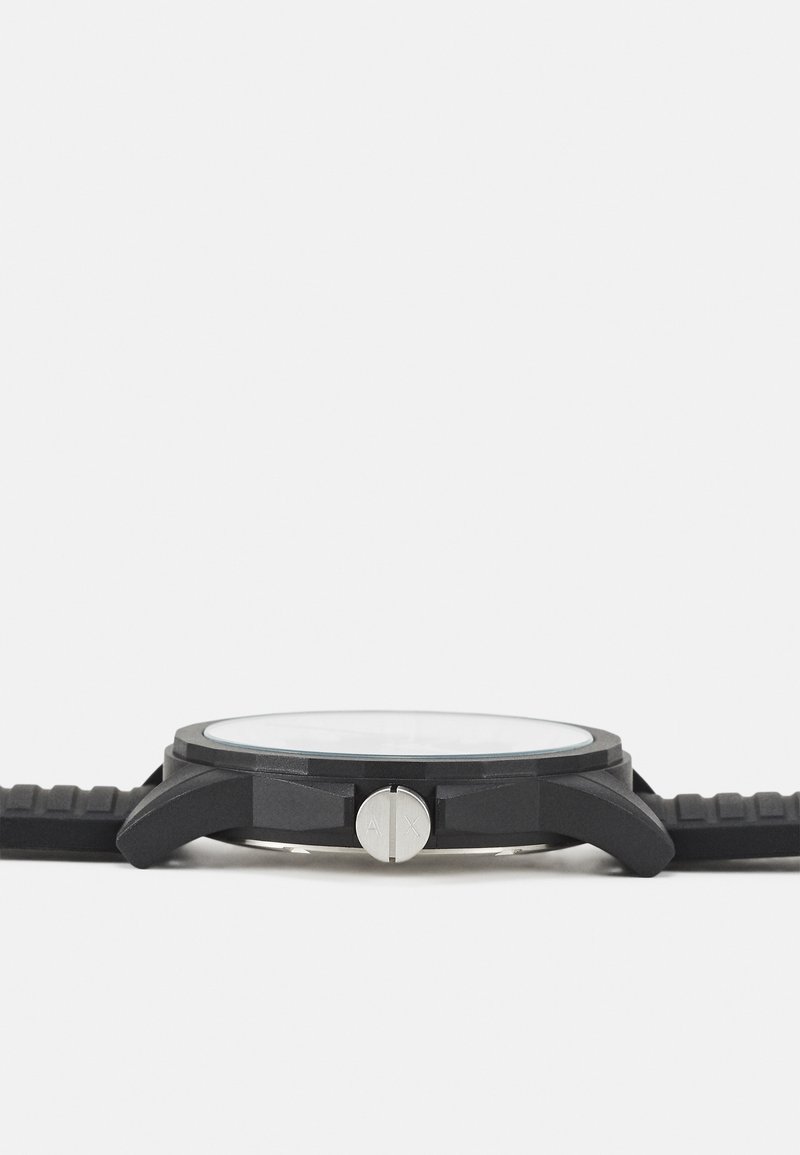 Reloj negro con caja redonda, pulsadores plateados y correa de silicona texturizada. La tapa de vidrio transparente revela una esfera minimalista.
