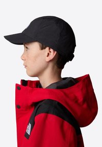 Chaqueta impermeable roja con detalles en negro, que cuenta con capucha y cierre de botones. Una gorra negra completa el atuendo, vista de lado.