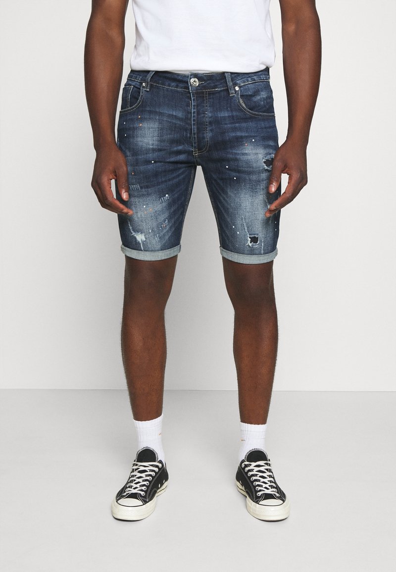 Shorts de mezclilla en azul oscuro con detalles desgastados, dobladillos enrollados y adornos. Combinados con zapatillas negras y calcetines blancos.