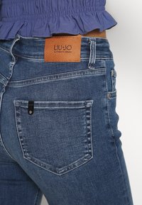 Tmavě modré džíny z denimového materiálu s texturovanou hnědou koženou štítkem na pase. Obsahují klasické zadní kapsy s malým černým štítkem.