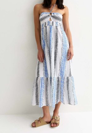 Kvinde iført en hvid midikjole med blå og sorte lodrette blomsterstriber, halterneck og beige sandaletter med spænde, stående op ad en ensfarvet væg.