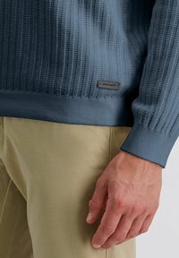 Helles blauer gerippter Pullover mit strukturiertem Stoff und einem einfarbigen Saum. Ein graues Etikett in der unteren linken Ecke zeigt den Markennamen "Pierre Cardin".