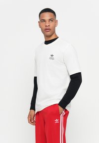 White short-sleeve t-shirt with black long sleeves underneath. Features a small black logo on the chest and relaxed fit. Red sweatpants with white stripes.