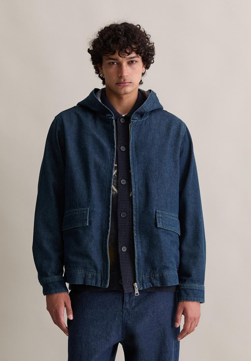 Dunkelblaue Jeansjacke mit Kapuze, Frontreißverschluss und zwei aufgesetzten Taschen. Verfügt über ein gefüttertes Innenfutter mit Reißverschluss, getragen über einem marineblauen Pullover.