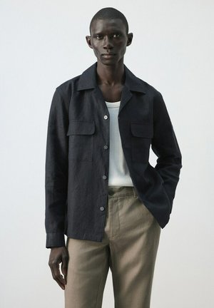 Hombre delgado con chaqueta negra abotonada sobre una camisa blanca y pantalones beige, de pie con una mano en el bolsillo contra un fondo liso.