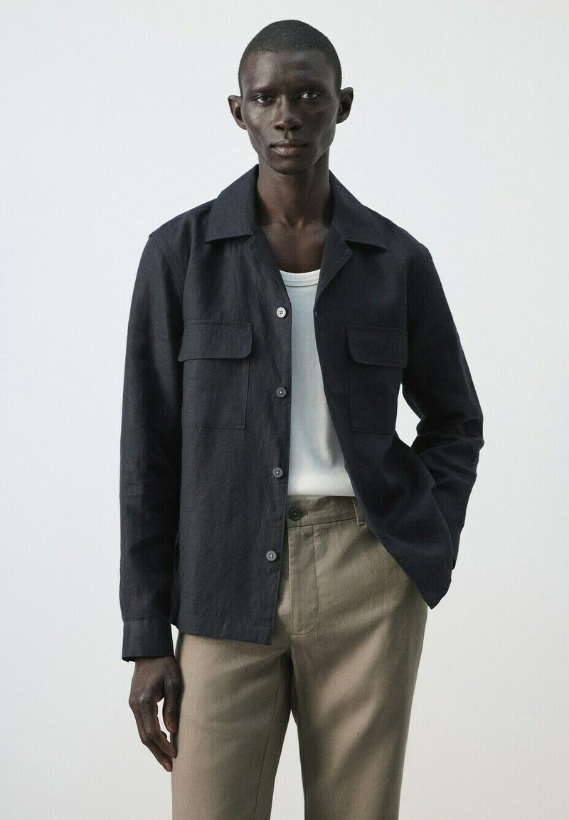 Homme élancé portant une veste noire boutonnée par-dessus une chemise blanche et un pantalon beige, debout, une main dans la poche, devant un fond uni.