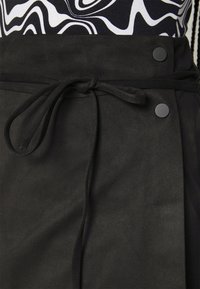 Svart nubuckomslutningskjol med bälte och cirkulära svarta knappar vid midjan, med en slät, strukturerad yta.