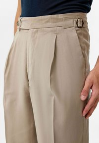 Beige geplooide broek met een hoge taille, voorzien van een zijgesp, een soepele textuur en een subtiel diagonale weefpatroon.