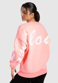 Sudadera de color rosa con texto en cursiva blanca en la parte posterior. Tiene un escote redondo, corte oversize y puños y dobladillo acanalados.