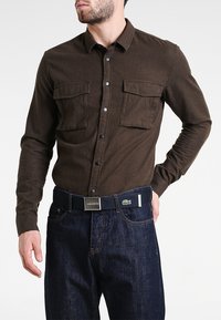 Brown long-sleeve button-up shirt with two chest pockets and black buttons, worn with dark denim jeans and a Lacoste belt.