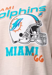 Lys beige T-shirt med Miami Dolphins-logo, tekst i teal og orange, samt grafik af en fodboldhjelm. Blødt bomuldsstof, afslappet pasform.