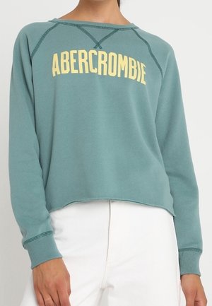 Bluza z długim rękawem w kolorze niebieskozielonym z żółtym napisem "ABERCROMBIE", noszona z białymi, wysokim stanem spodniami, widoczna krótka część środkowa.