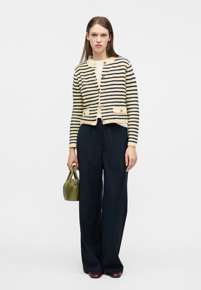 Γυναίκα που φοράει κρεμ και ναυτικό μπλε ριγωτή ζακέτα με κουμπιά, ναυτικά παντελόνια φαρδιάς γραμμής, καφέ παπούτσια και κρατάει μια μικρή τσάντα ελιάς.