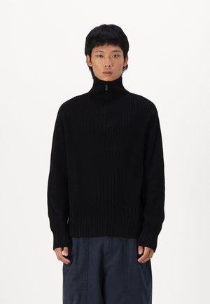 Maglione a coste nere con colletto alto e chiusura a mezza zip. Presenta maniche lunghe e una vestibilità morbida, abbinato a pantaloni blu navy.
